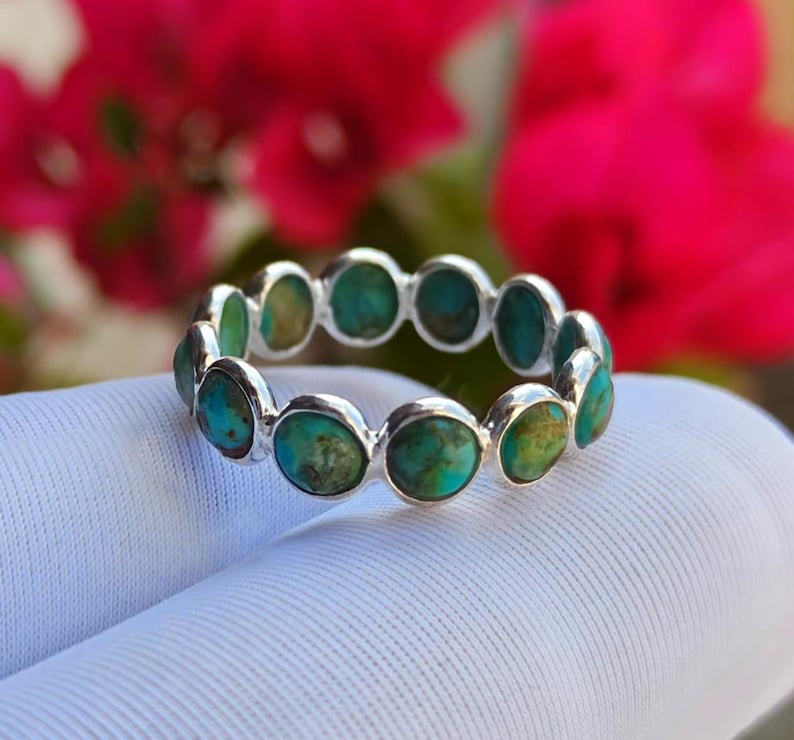 May include: A silver ring set with turquoise stones. The ring has a repeating pattern of round turquoise stones, each set in a silver bezel. The background features blurred pink flowers, with the ring resting on a white surface.