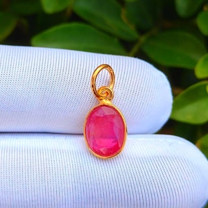 Könnte beinhalten: Goldener Anhänger mit einem ovalen, facettierten, leuchtend rosa Edelstein. Der Anhänger hat eine kleine Schlaufe oben für eine Kette. Der Edelstein ist in einer goldenen Fassung gefasst.