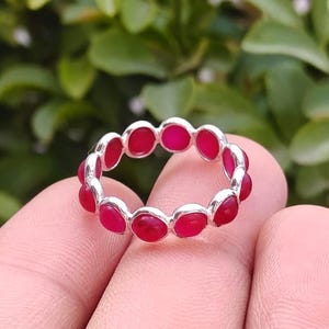 Può includere: Un anello d'argento con una fascia continua di pietre preziose rosse brillanti di forma ovale. Il design dell'anello presenta un motivo ripetuto di pietre rosse, creando un gioiello audace. Le pietre sono molto vicine.