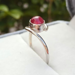 Puede incluir: Un anillo de plata con una vibrante gema roja. La gema redonda está engastada en un bisel de plata. La banda del anillo tiene un diseño curvo, creando un diseño elegante y llamativo. Una joya.