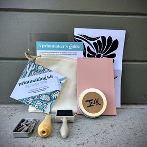 May include: Printmaking kit with tools, ink, and a guide. Includes a roller, carving tools, and a tin of ink. The kit also contains a canvas bag and a pink card. The guide is titled "A printmaker's guide".