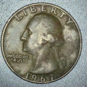 May include: A silver quarter coin from 1967, featuring a profile of George Washington and the inscriptions "LIBERTY" and "IN GOD WE TRUST." The coin has a worn appearance.