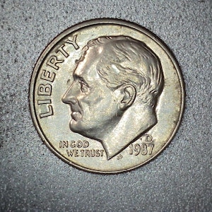 May include: A close-up of a silver dime coin. The coin features a profile of a man's head, the word "LIBERTY", and the phrase "IN GOD WE TRUST". The year "1987" is visible. The coin is on a textured gray surface.