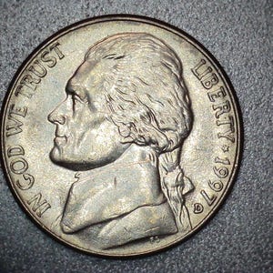 May include: A silver 1997 nickel coin featuring a profile of Thomas Jefferson. The coin has the inscriptions "IN GOD WE TRUST" and "LIBERTY" along the rim. The coin is on a dark gray surface.