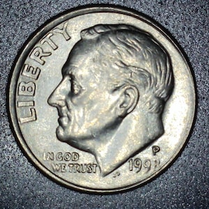 Op de afbeelding: Een close-up van een Amerikaanse dime-munt. De munt toont een profiel van een man, het woord "LIBERTY" en de zin "IN GOD WE TRUST". Het jaartal "1991" is zichtbaar. De munt is zilverkleurig.