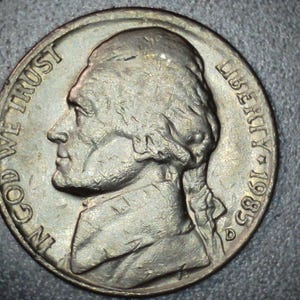 May include: A close-up of a United States nickel coin. The coin features a profile of Thomas Jefferson, the words "IN GOD WE TRUST" and "LIBERTY", and the year 1985. The coin is silver in color.