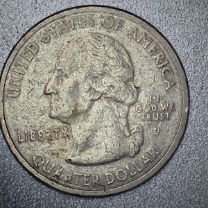 May include: A United States quarter coin, featuring a profile of George Washington, the words "Liberty," "Quarter Dollar," and "In God We Trust." The coin is silver in color and has a slightly worn appearance.