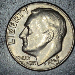 May include: A close-up of a silver dime coin. The coin features a profile of a man, the word "LIBERTY" at the top, and the words "IN GOD WE TRUST" and the date "1975" at the bottom. The coin is on a dark surface.