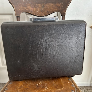 Puede incluir: Un maletín rectangular de color marrón oscuro con un acabado texturizado similar al cuero. El maletín cuenta con un asa de metal plateado y un borde plateado a juego en la parte superior que sirve como mecanismo de cierre seguro.