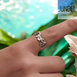 Può includere: Un anello color argento con un motivo ripetuto di piccole figure di orso. L'anello è indossato su un dito, con uno sfondo sfocato di acqua e fogliame. La parola "JUNDEL" è visibile nell'angolo in alto a destra.