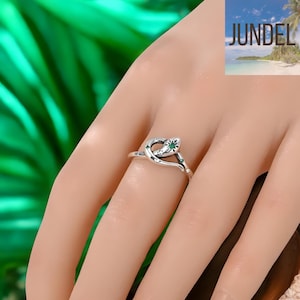 May include: A silver-toned ring in the shape of a snake, with a green gemstone eye and smaller green gems along the band. The ring is worn on a finger, with the word "JUNDEL" visible in the background.