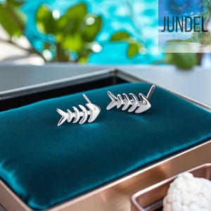 May include: Silver-coloured fishbone earrings. Each earring has a fish skeleton design with a small, round accent. The earrings are presented on a teal velvet surface, with the brand name "JUNDEL" visible in the background.