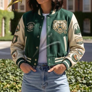 May include: A dark green and cream varsity jacket featuring a white undershirt and blue jeans. The jacket has a bear head embroidery, a compass graphic with the text Adventure Awaits, and various nature-themed patches on the sleeves.