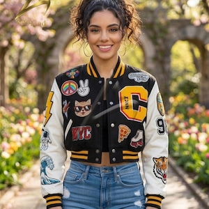 May include: A black and white varsity jacket featuring yellow striped ribbed cuffs and collar. The jacket is adorned with various embroidered patches including a large yellow letter C, a cat with sunglasses, a peace sign, a pizza slice, and a tiger.