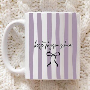 Könnte beinhalten: Weiße Keramik-Tasse mit vertikalen lavendelfarbenen Streifen und dem Text "beste physio silvia" in schwarzer Schrift. Unter dem Text befindet sich eine schwarze Schleifen-Grafik. Die Tasse hat einen weißen Henkel und steht auf einer weißen Strickoberfläche.