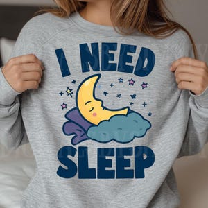 May include: A light gray sweatshirt with the words "I NEED SLEEP" in navy blue block letters. The design includes a yellow crescent moon with closed eyes, resting on a blue cloud and pillow, surrounded by stars.