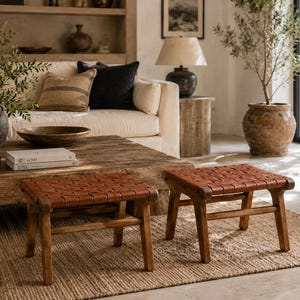 May include: Two low stools featuring dark brown wooden frames and seats constructed from woven leather strips. The stools have four legs connected by horizontal support bars, providing a stable base for seating in a living space.
