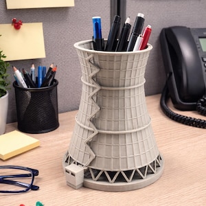 May include: A light gray, 3D-printed desk organizer shaped like a nuclear cooling tower. The cylindrical structure features a grid pattern, horizontal rings, and a small attached building at the base. It holds various pens and pencils upright.