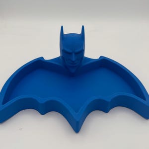 May include: A bright blue, bat-shaped tray featuring a sculpted, three-dimensional head of a masked character with pointed ears at the center. The tray has a recessed, wing-shaped basin with raised edges, providing a flat surface for holding small items.