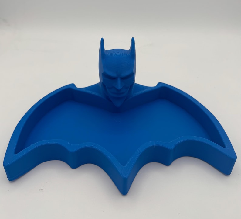 May include: A bright blue, bat-shaped tray featuring a sculpted, three-dimensional head of a masked character with pointed ears at the center. The tray has a recessed, wing-shaped basin with raised edges, providing a flat surface for holding small items.