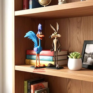 May include: Two standing cartoon character figurines on a wooden shelf. On the left, a blue and orange Road Runner bird figure. On the right, a brown and tan Wile E. Coyote figure. Both figures are mounted on black rectangular bases.