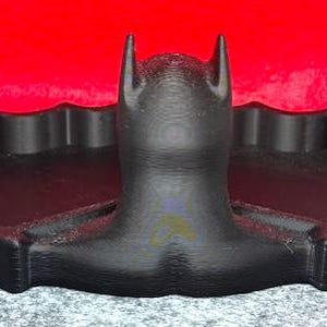 May include: A black, 3D-printed tray shaped like a bat silhouette. The central portion features a raised, stylized head with pointed ears, flanked by two recessed, wing-shaped compartments for storage. The surface has a visible, layered texture.