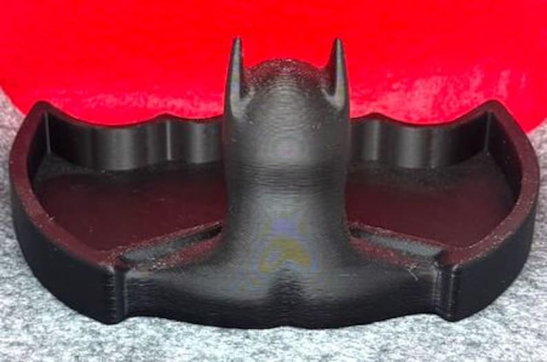 May include: A black, 3D-printed tray shaped like a bat silhouette. The central portion features a raised, stylized head with pointed ears, flanked by two recessed, wing-shaped compartments for storage. The surface has a visible, layered texture.