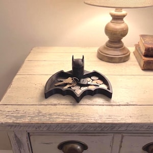 May include: A black, bat-shaped tray featuring a central sculpted head figure. The tray is filled with various metal keys and assorted circular coins. The design is symmetrical with pointed wing-like edges extending from the central figure.