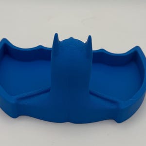 May include: A solid blue 3D printed tray shaped like a stylized bat silhouette. The object features a central raised head section with pointed ears and two symmetrical recessed wing-shaped compartments on either side for holding small items.