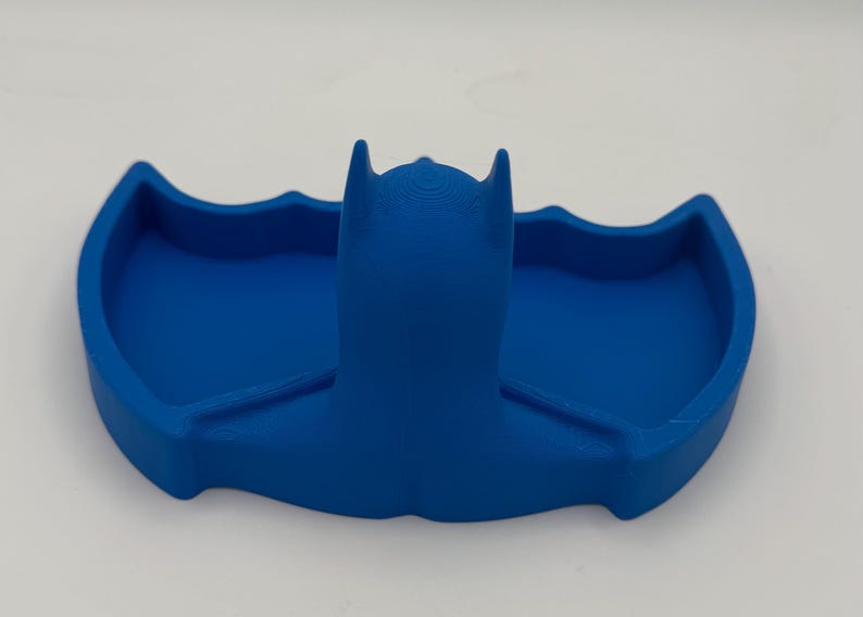 May include: A solid blue 3D printed tray shaped like a stylized bat silhouette. The object features a central raised head section with pointed ears and two symmetrical recessed wing-shaped compartments on either side for holding small items.
