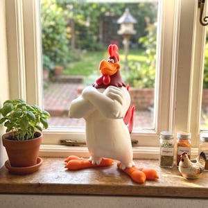 May include: A cartoon-style rooster figurine with a white body, red comb, and orange beak and feet, standing with its wings crossed over its chest. The figure has a smooth, matte finish and is positioned in a confident, upright posture.