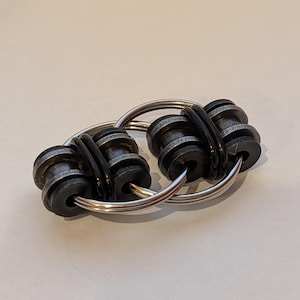 Puede incluir: Un juguete fidget de metal plateado con dos anillos entrelazados. Cada anillo tiene dos juegos de cuentas cilíndricas negras. Las cuentas están separadas por anillos de goma negros. El juguete está diseñado para aliviar el estrés y la concentración.