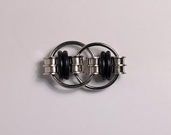 May include: A silver metal fidget toy with two interlocking rings. Each ring has two black rubber bands and silver metal cylinders. The toy is designed to be manipulated and provide sensory stimulation.