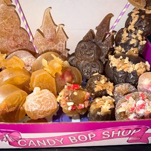 Könnte beinhalten: Eine Schachtel mit der Aufschrift CANDY BOP SHOP enthält eine Auswahl an Halloween-Duftwachsen oder Seifen. Die Kollektion umfasst bernsteinfarbene und dunkelbraune Formen wie Kürbisse, Fledermäuse, Schlösser und Kürbisgesichter.
