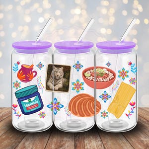 Può includere: Tre bicchieri di vetro trasparente con coperchi e cannucce viola. Ogni bicchiere presenta adesivi colorati di un vaso, una tigre, una ciotola di cibo, una pasticceria, un tamale e un barattolo di VapoRub. Disegni floreali e geometrici decorano i bicchieri.