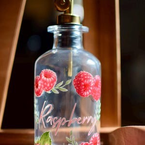 May include: Clear glass bottle with a gold spout, decorated with red raspberry illustrations and the word "Raspberry" in pink script. The bottle is designed for dispensing liquids.