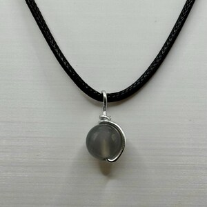 May include: A gray gemstone pendant necklace. The pendant is a round, polished gray stone, wrapped in silver-colored wire, and suspended from a black cord. The background is a light gray, textured surface.