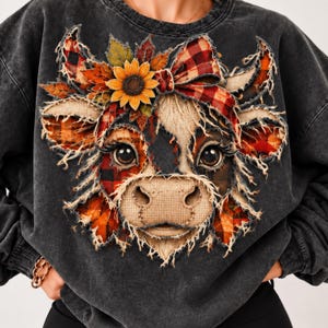 Op de afbeelding: Een antracietkleurige acid-wash sweater met een centrale afbeelding van een koeienkop. De koe is versierd met een rood-zwarte geruite strik, een gele zonnebloem en herfstbladeren, allemaal weergegeven met een gerafeld, textuurrijk effect.