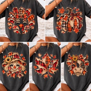 May include: A charcoal gray t-shirt featuring five distinct fall-themed graphic prints: a turkey, a pumpkin, a scarecrow, a maple leaf, and a cow, all decorated with frayed-edge red and black plaid patterns and autumn-colored accents.