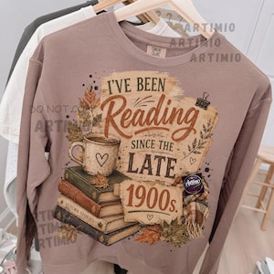 May include: A mauve crewneck sweatshirt featuring a graphic print of a stack of books, a coffee mug with a heart, autumn leaves, and a plaid scarf. The text reads I'VE BEEN Reading SINCE THE LATE 1900s. The design includes a paper-like background.