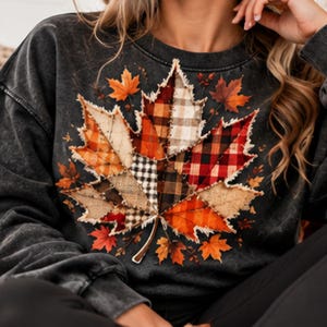 Op de afbeelding: Een antracietgrijs sweatshirt met ronde hals en een grote esdoornblad-afbeelding in het midden. Het blad is samengesteld uit diverse stofachtige patches met ruitpatronen in oranje, rood, bruin en crème, voorzien van gerafelde randen.