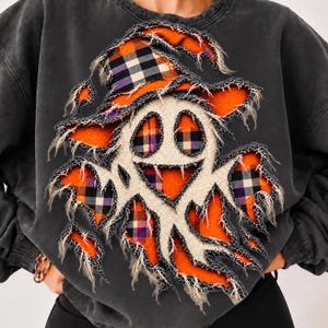 May include: A charcoal gray crewneck sweatshirt featuring a distressed-style applique of a ghost wearing a witch hat. The ghost and hat are constructed from layers of orange fabric and a black, white, and purple plaid pattern with frayed edges.