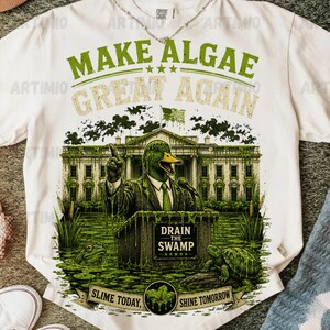 Könnte beinhalten: Ein cremefarbenes T-Shirt mit einer Grafik, die eine Ente im Anzug an einem Rednerpult vor einem Gebäude zeigt. Der Text lautet "MAKE ALGAE GREAT AGAIN", "DRAIN THE SWAMP" und "SLIME TODAY, SHINE TOMORROW". Ein Frosch und eine Schildkröte sind abgebildet.