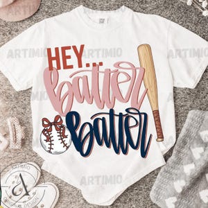 Può includere: T-shirt bianco con la scritta "HEY... batter batter" in rosa e blu navy. Una mazza da baseball e una palla da baseball con un fiocco rosso sono presenti nel design. La maglietta è in stile casual.