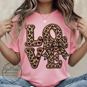 May include: A pink t-shirt with the word "LOVE" in a leopard print design and heart accents. The "O" is a bow. The shirt also has the text "AjaNess DIGITAL DESIGNS" in a circle at the bottom left.