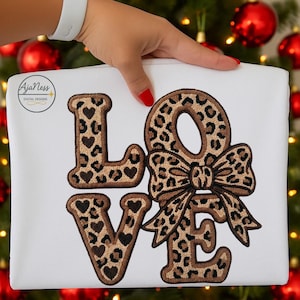 May include: White t-shirt featuring the word "LOVE" embroidered in a leopard print pattern. The letters "L" and "E" have heart-shaped details. The "O" is a bow. The t-shirt is held by a person with a white wristband and red nail polish.