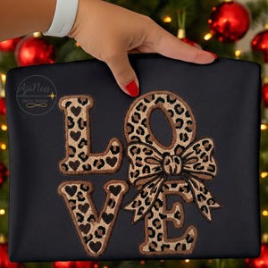 May include: Black fabric featuring the word "LOVE" embroidered in a leopard print pattern. The letters "L" and "V" have heart details. The "O" is designed as a bow. The text "AjaNess DIGITAL DESIGNS" is in the upper left corner.