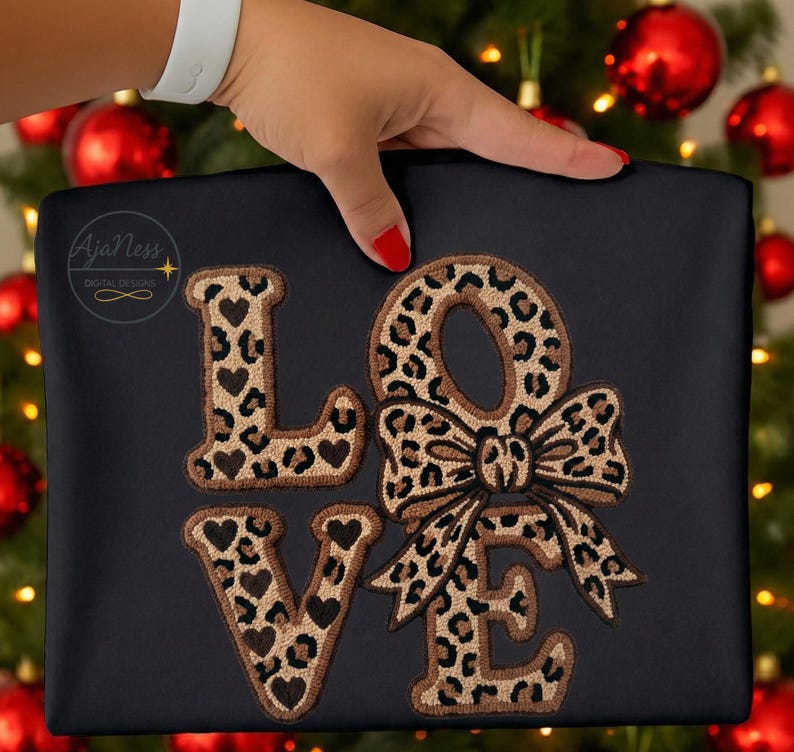 May include: Black fabric featuring the word "LOVE" embroidered in a leopard print pattern. The letters "L" and "V" have heart details. The "O" is designed as a bow. The text "AjaNess DIGITAL DESIGNS" is in the upper left corner.
