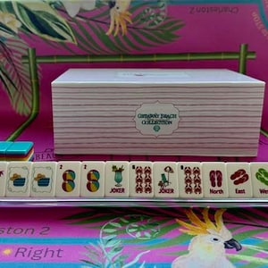 May include: A rectangular white box with horizontal stripes and a label reading Getaway Beach Collection, positioned behind a row of mahjong tiles on a clear rack. The tiles feature colorful beach-themed icons including palm trees, beach balls, cocktails, crabs, and flip-flops labeled North, East, West, and South.