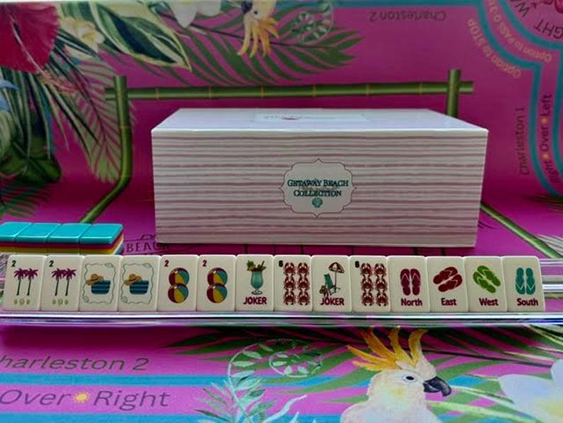 May include: A rectangular white box with horizontal stripes and a label reading Getaway Beach Collection, positioned behind a row of mahjong tiles on a clear rack. The tiles feature colorful beach-themed icons including palm trees, beach balls, cocktails, crabs, and flip-flops labeled North, East, West, and South.